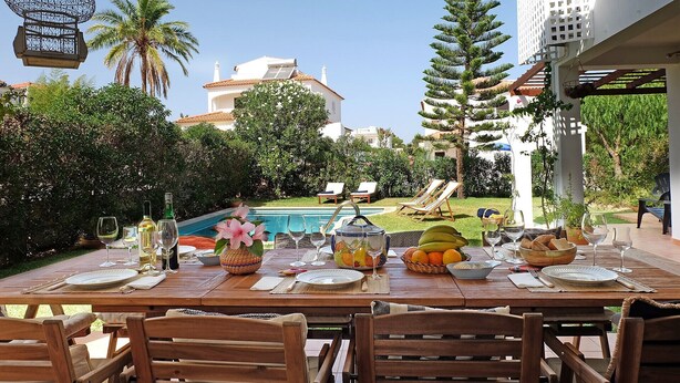 An outdoor dining area with a pool, lounge chairs, and a view of the surrounding greenery and buildings. - VV Beautiful Villa w/pool | A/C | VILAMOURA! (Quarteira)