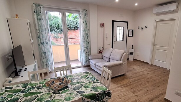 Ein Wohnzimmer mit einer Couch, einem Esstisch mit Tischdecke mit Blätermotiv, einem Fernseher auf einem Schrank und einer Glasschiebetür, die zu einem Außenbereich führt.
