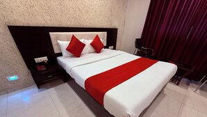 A neatly made bed with a white and red bedspread, a dark headboard, and a red accent pillow. There is a nightstand with a phone and a lamp, and a red curtain in the background. - Hotel Arya Residency Rabale (Navi Mumbai)