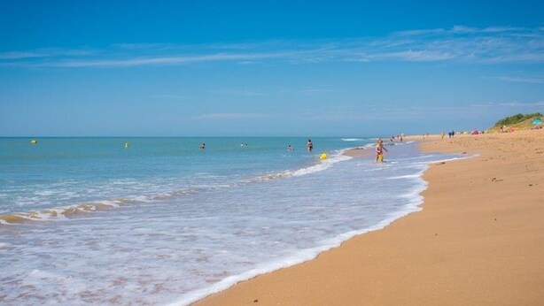 Une vaste plage de sable fin, avec de douces vagues, un ciel bleu clair et, au loin, des gens qui profitent de l’eau.