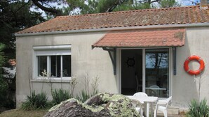 Una pequeña casa con techo de tejas, una puerta blanca y una ventana. Hay una mesa blanca y una silla blanca afuera.