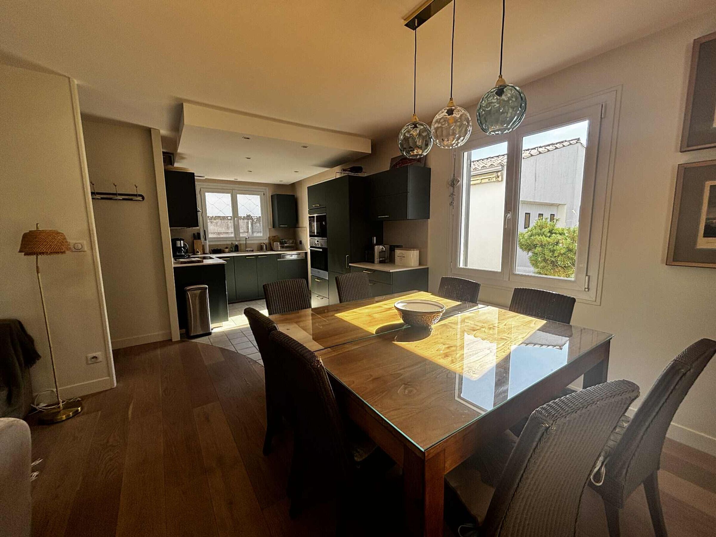 A modern kitchen with dark cabinets, a dining area with a glass table, and a view of the outside through a large window.
