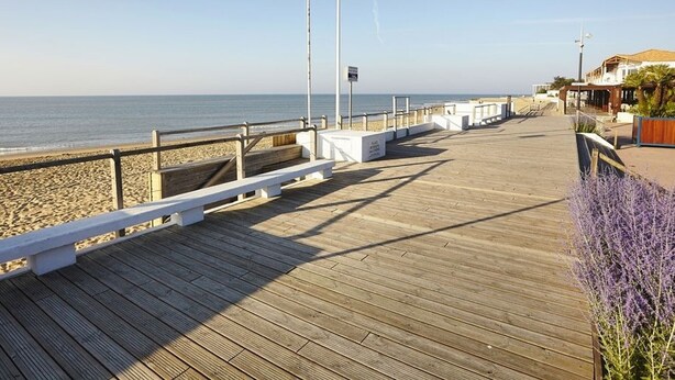 A wooden boardwalk leading to a beach with a flag, benches, and a building with a balcony.