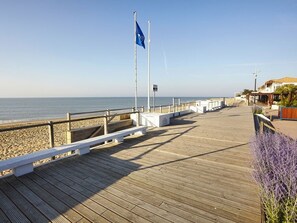 En träpromenad som leder till en strand med en flagga, bänkar och en byggnad med en balkong.