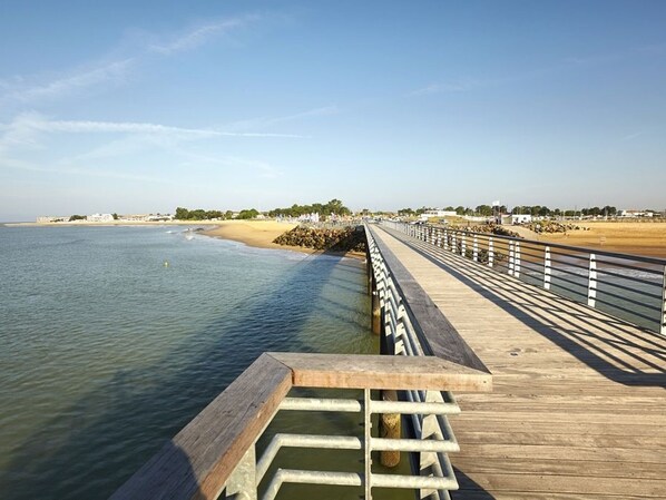 Un muelle de madera se extiende sobre aguas tranquilas, con una playa de arena y edificios lejanos en la orilla.