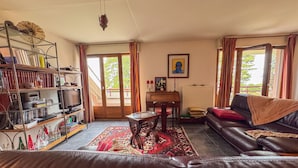 A living room with a leather sofa, a wooden coffee table, a bookshelf, and a dining table with chairs. There is a large window with curtains and a view of greenery outside. - Duplex with Terrace and Private Beach Access, Garage Included (Évian-les-Bains)