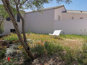 Un bâtiment blanc avec un toit en pente, un banc blanc et un jardin où poussent différentes plantes et fleurs.