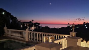 Uma varanda com piscina, com vista para a área costeira ao crepúsculo, com a lua visível.