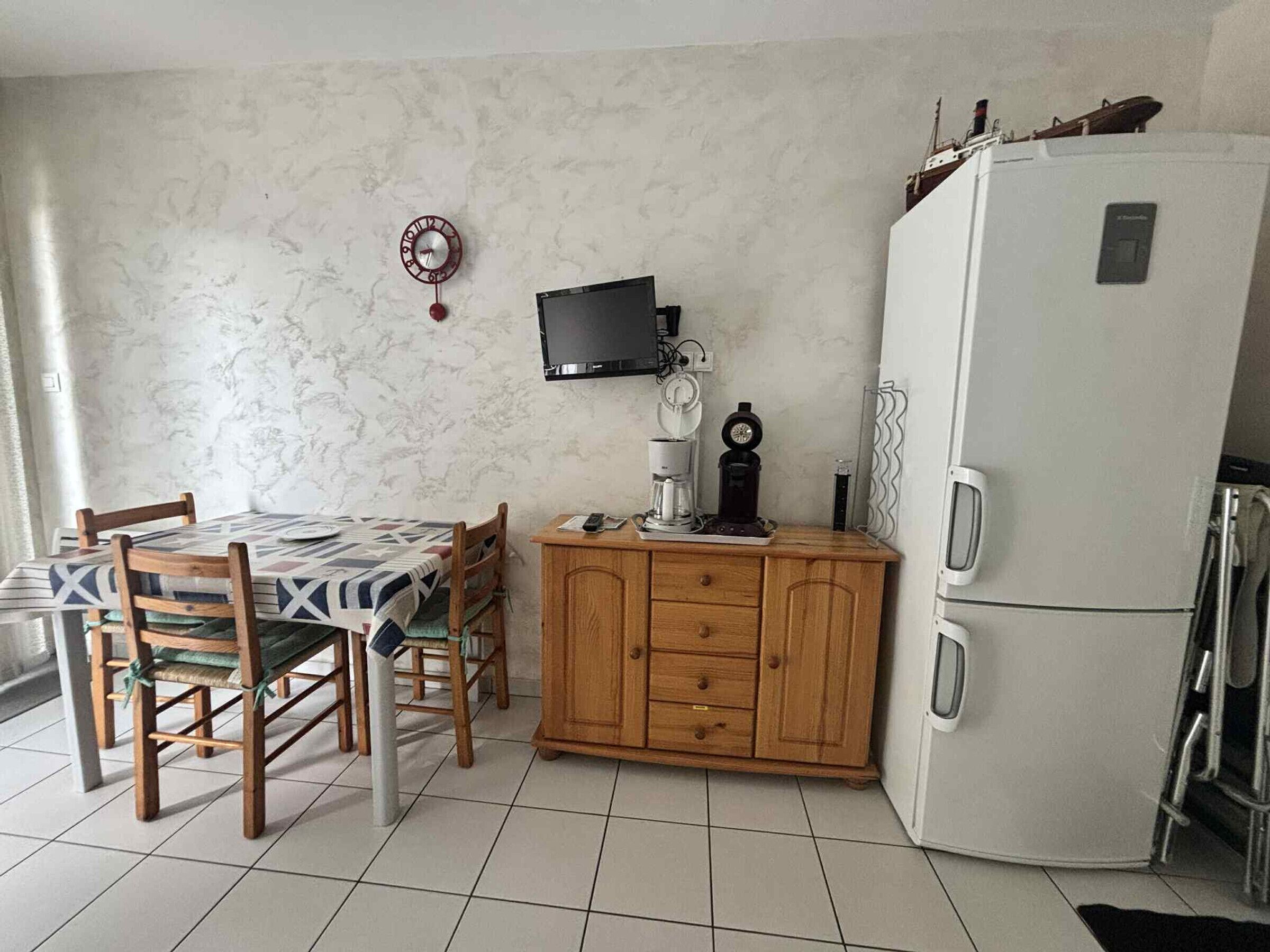 Une cuisine équipée d’une table à manger, de chaises, d’un réfrigérateur, d’une machine à café et d’une horloge murale.