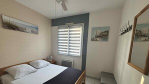 A bedroom with a bed, a ceiling fan, a window with blinds, and artwork on the walls. - Cozy city-center apartment, 400m from beaches, parking included (La Tranche-sur-Mer)