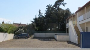 A parking area with a car, a building with a staircase, and trees in the background. - Cozy city-center apartment, 400m from beaches, parking included (La Tranche-sur-Mer)