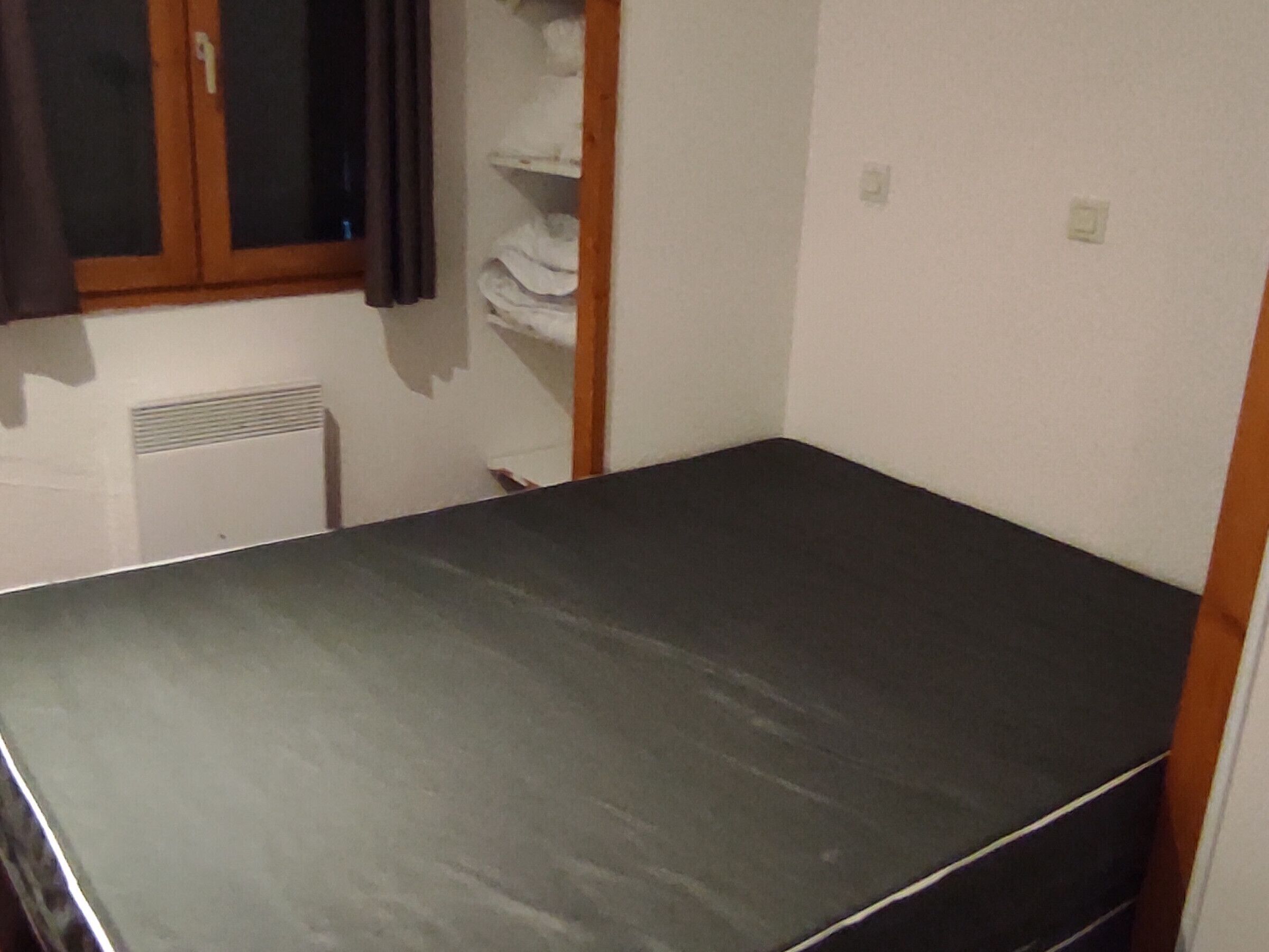 Una habitación pequeña y tenuemente iluminada, con una cama individual, un radiador y una ventana con cortinas.