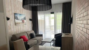 A modern hotel room with a wooden accent wall, a grey sofa with red and black cushions, a glass-top dining table, a black chair, and a small wooden side table. - Studio in Gourette with pool and pets allowed, 4 pers (Gourette)