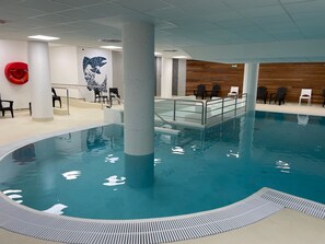 Piscina interior com superfície de água transparente, rodeada por um deck com guarda-corpo de metal. Há cadeiras e um salva-vidas no deck.