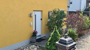 En gul byggnad med en dörr, en liten trädgård med växter och en dekorativ stenkonstruktion.