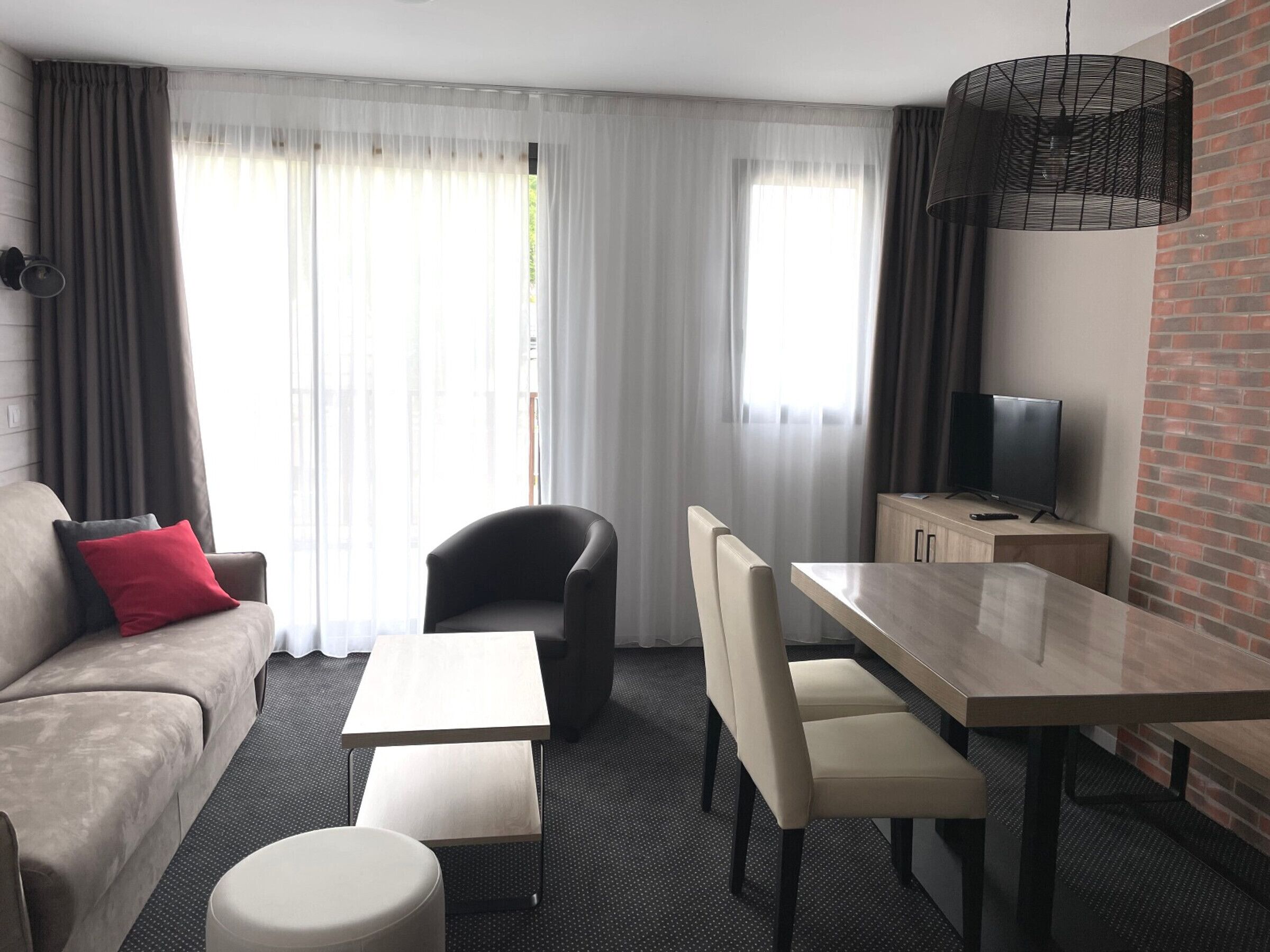 A modern hotel room with a sofa, a dining table, chairs, a TV, and a brick accent wall.