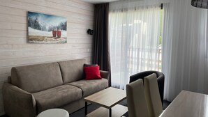 Een moderne hotelkamer met een bank, een klein tafeltje en een stoel. Er hangt een schilderij aan de muur en er is een raam met gordijnen.