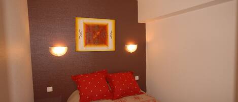 A hotel room with a bed, red pillows, a wall with a framed picture, and two wall-mounted lights.