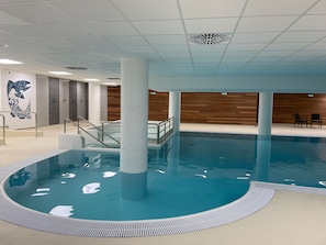 En indendørs swimmingpool med en central søjle, trapper der fører ned i poolen, og en trævæg i baggrunden.