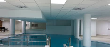 An indoor swimming pool with clear water, surrounded by white walls and columns, and a tiled ceiling with ventilation grilles.