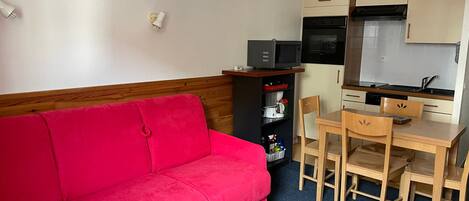 A compact living space with a red sofa, a wooden dining table with four chairs, a kitchenette with a microwave, oven, and sink, and a small shelf with kitchen items.