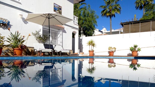 Ein Swimmingpool mit Spiegelung, Topfpflanzen, ein weißes Gebäude und eine Palmenbaum.