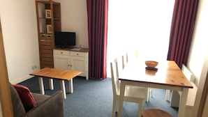 A dining area with a wooden table, white chairs, a small wooden bench, a television on a cabinet, and red curtains. - Comfortable studio 6 pers, slopes view, pets allowed (Gourette)
