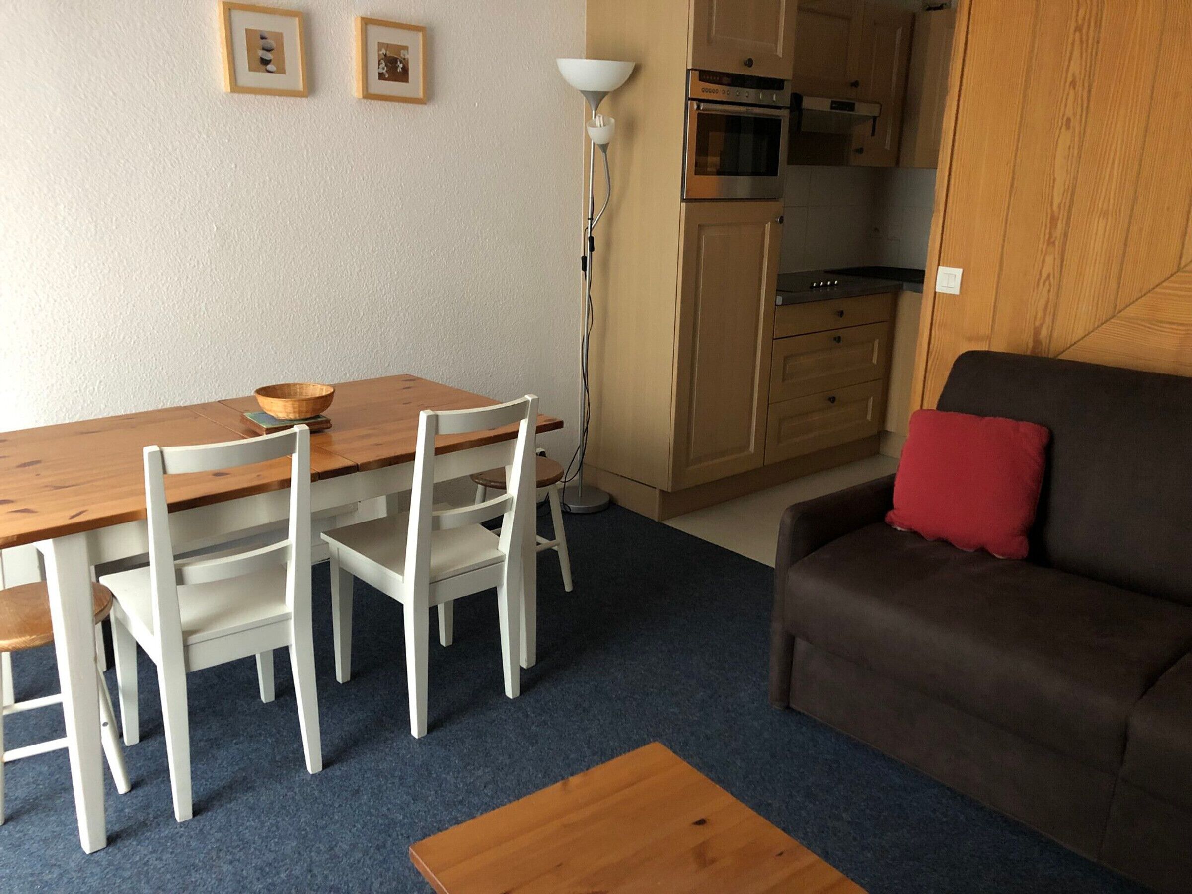 A compact living space with a wooden dining table, white chairs, a brown sofa with a red pillow, and a kitchenette in the background.