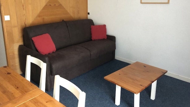 A room with a wooden partition, a brown sofa with red cushions, a white chair, and a small wooden table. - Comfortable studio 6 pers, slopes view, pets allowed (Gourette)
