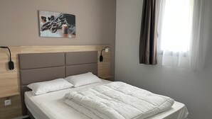 Ein ordentlich bezogenes Bett mit weißer Decke und Kissen, ein Holz-Kopfteil, ein Wandbild und ein Fenster mit Vorhängen.
