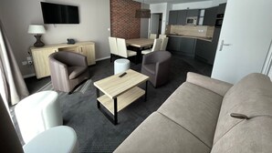 Uma sala de estar moderna com um sofá, poltronas, uma mesa de centro e uma televisão. É visível uma cozinha ao fundo, com um micro-ondas e uma pia.