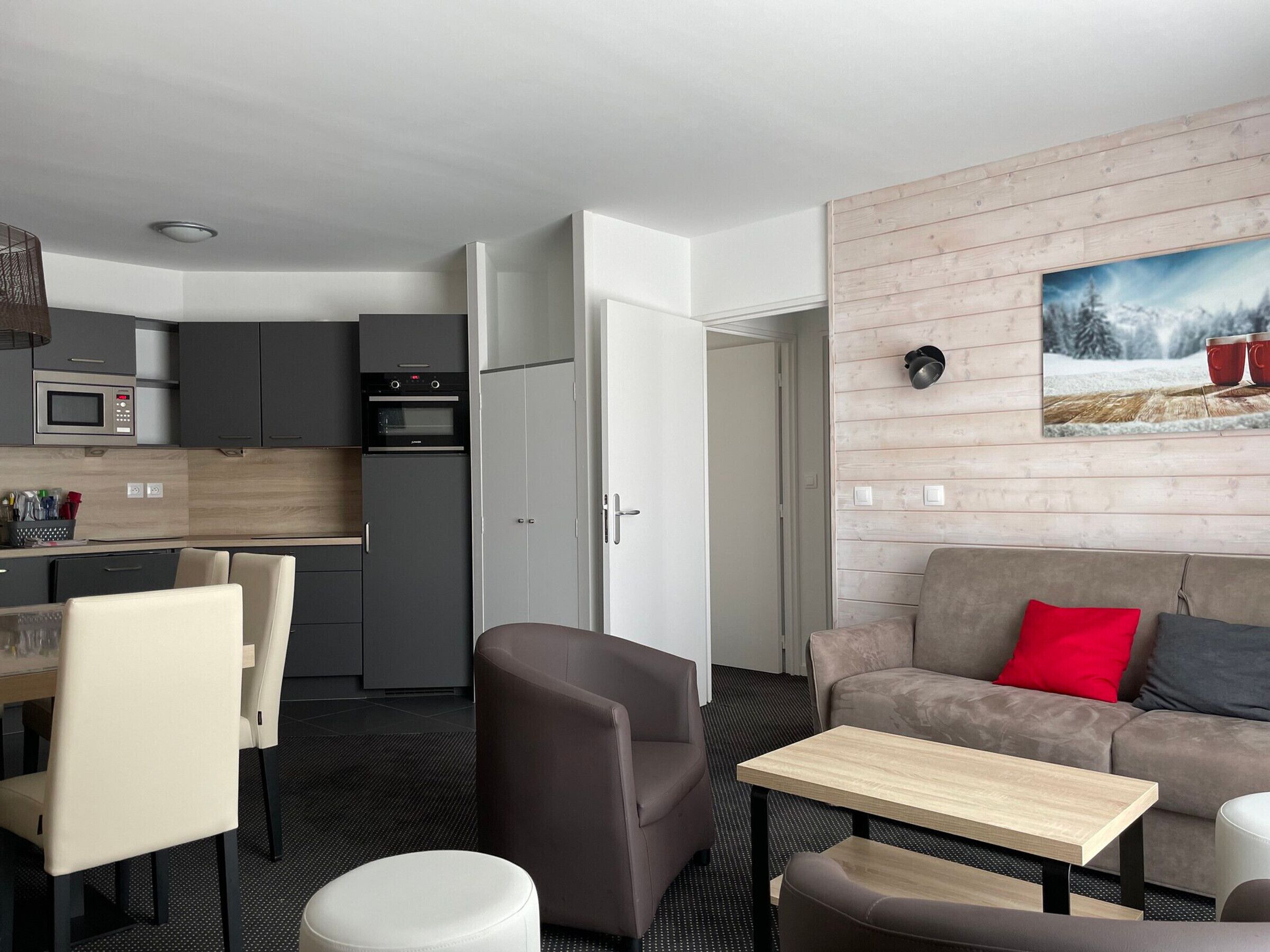 A modern kitchen with grey cabinets and a microwave, a dining area with a table and chairs, and a living area with a sofa and a painting of a snowy landscape.