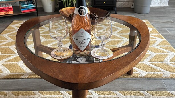 A round wooden table with a glass top, holding a bottle of Pinot Grigio sparkling wine, two wine glasses, and a decorative item. - Excellent 2 bed cottage in the stunning Scottish Highlands near Portmahomack (Tain)
