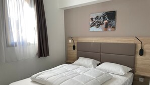 Una cama bien hecha con ropa de cama blanca, un cabecero con acabado en madera clara y un cuadro colgado en la pared que representa un paisaje invernal.