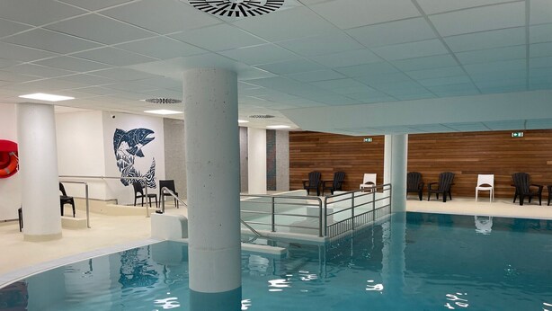 Una piscina interior con agua cristalina, rodeada de sillas de descanso y un muro de madera.