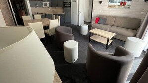 Ett modernt vardagsrum med en soffa, ett soffbord och stolar. Det finns en matplats med ett bord och stolar. Köket syns i bakgrunden, med vitvaror och skåp.