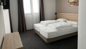 Et hotelværelse med seng, skrivebord, lampe, et billede på væggen og et vindue med gardiner.