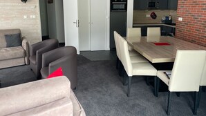 En moderne stue med sofa, stole og et spisebord. Der er et køkkenområde med hårde hvidevarer og en murstensvæg.