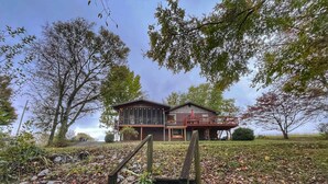 A two-story wooden house with a balcony, surrounded by trees and a grassy area with fallen leaves. - Sunfish Cove – Pet-Friendly Lakeside Retreat (Scottsville)