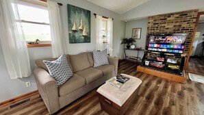 A living room with a beige sofa, patterned cushions, a coffee table, a TV stand with a video game console, and a brick-patterned wall. - Sunfish Cove – Pet-Friendly Lakeside Retreat (Scottsville)