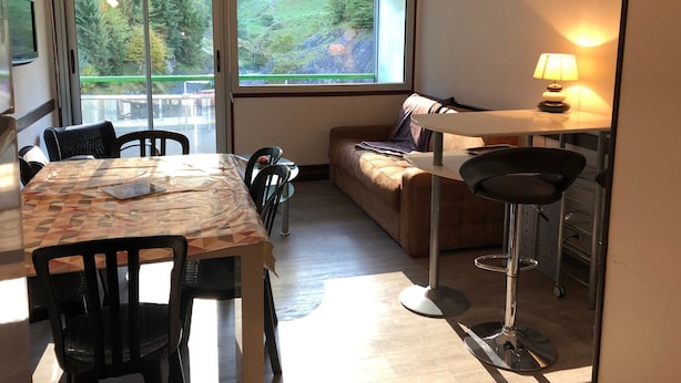 A dining area with a table and chairs, a sofa, a lamp, and a view of greenery through large windows. - Gourette Studio, 6 pax, WIFI, pets allowed (Gourette)