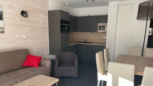 Un salon moderne avec un canapé, une petite table et un fauteuil gris. On aperçoit à l’arrière-plan un coin repas avec une table et des chaises.
