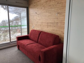A red sofa in a room with wooden panel walls and a large window offering a view of a snowy landscape. - Comfortable studio, 6 pers, mountain view, pets allowed, sheets provided (Gourette)