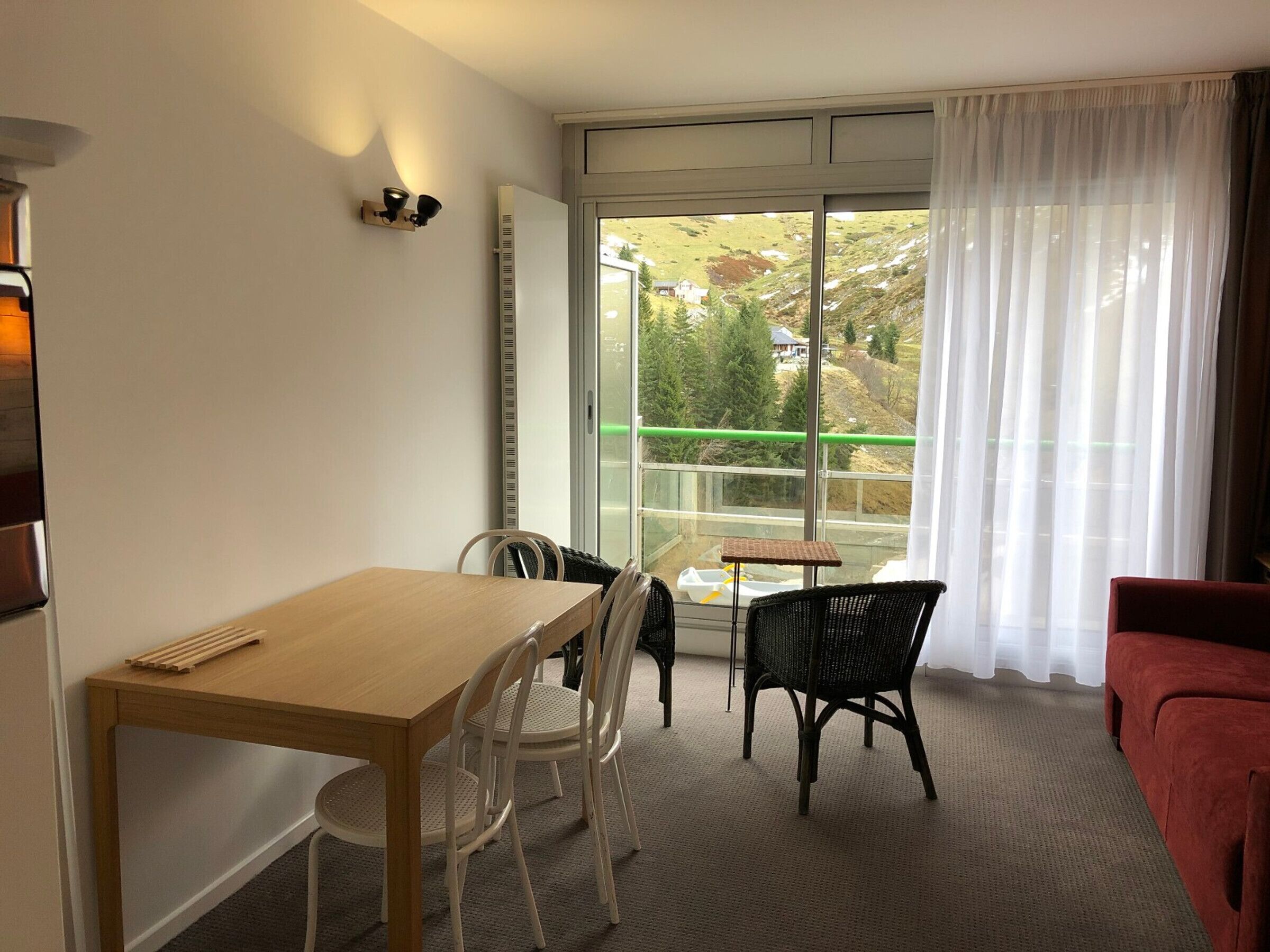 Una habitación con una mesa y sillas de madera, un sofá rojo y un ventanal con vistas a árboles y una casa.