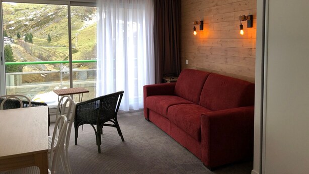 A room with a red sofa, a dining table with chairs, a window with curtains, and a view of a hillside. - Comfortable studio, 6 pers, mountain view, pets allowed, sheets provided (Gourette)
