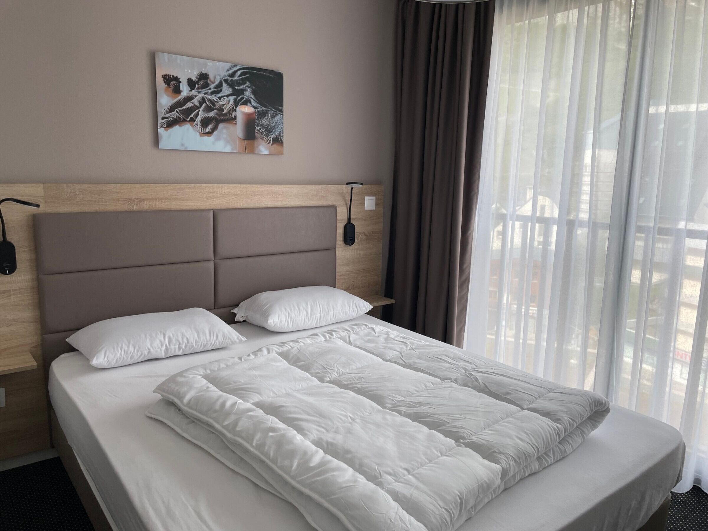 Ein Hotelzimmer mit einem großen Bett, einem Kopfteil, einem Nachttisch, einer Lampe, einem Bild an der Wand und einem Fenster mit Vorhängen.