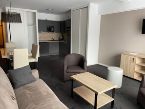 A modern hotel room with a sofa, a small table, a chair, a desk, a lamp, a TV, and a kitchenette in the background. - Studio cabin Gourette 6 ppl with pool and pets allowed (Gourette)