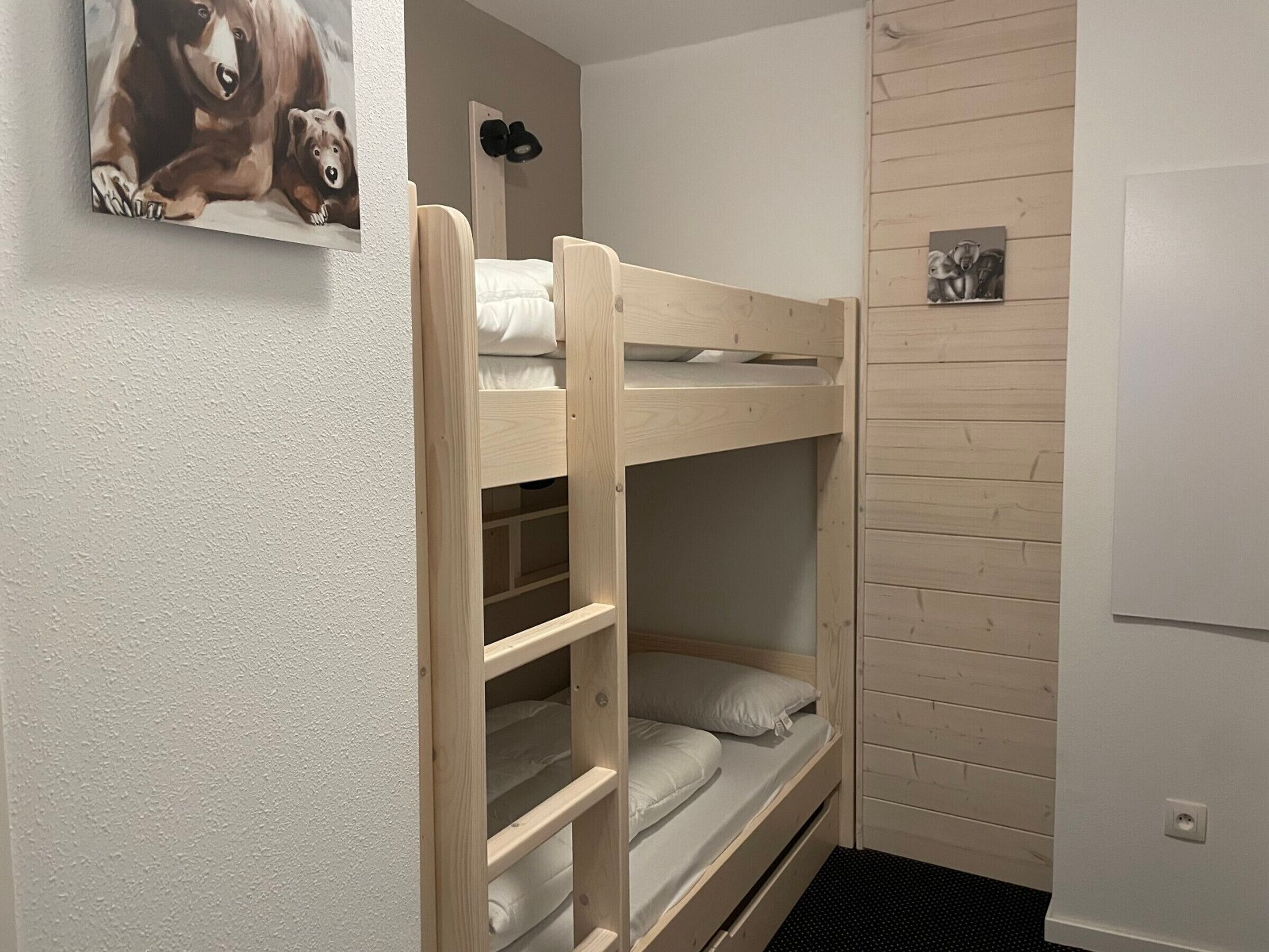 Ett rum med en våningssäng, ett träfärgat sänggavel, en liten hylla och en tavla som hänger på väggen.