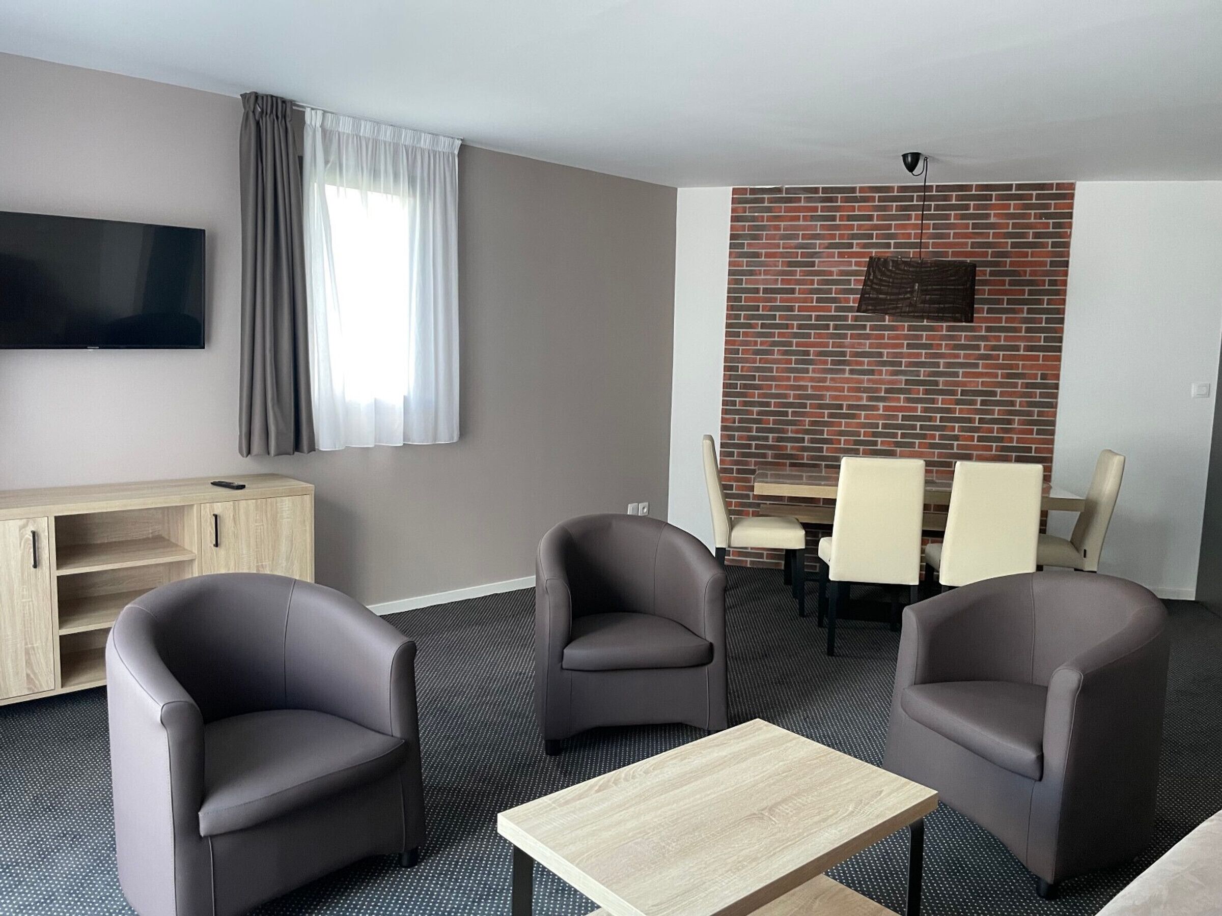 A modern hotel room with a flat-screen TV, a wooden cabinet, two armchairs, a small table, a brick accent wall, and a dining area with chairs.