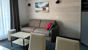 Una sala de estar moderna con un sofá, dos sillones, una mesita y un cuadro de un paisaje nevado, además de una zona de cocina.